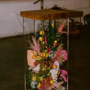 A transparent display case with a wooden top containing an arrangement of dried flowers, fruits, and decorative elements on a floor with a Acrylic Cocktail Table in the background.