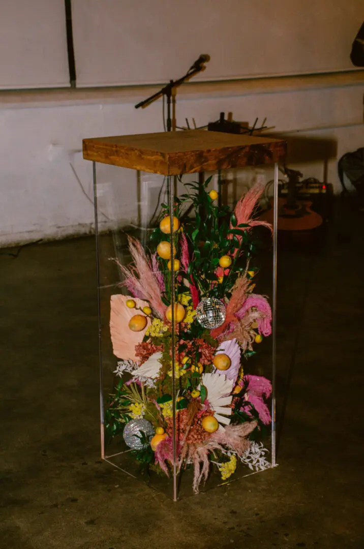 A transparent display case with a wooden top containing an arrangement of dried flowers, fruits, and decorative elements on a floor with a Acrylic Cocktail Table in the background.