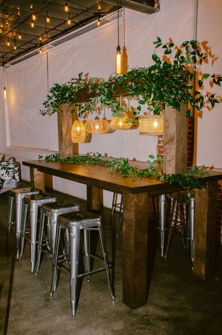 A cozy bar setting with a Rustic Kings Table, metal stools, and decorative greenery under warm pendant lighting.