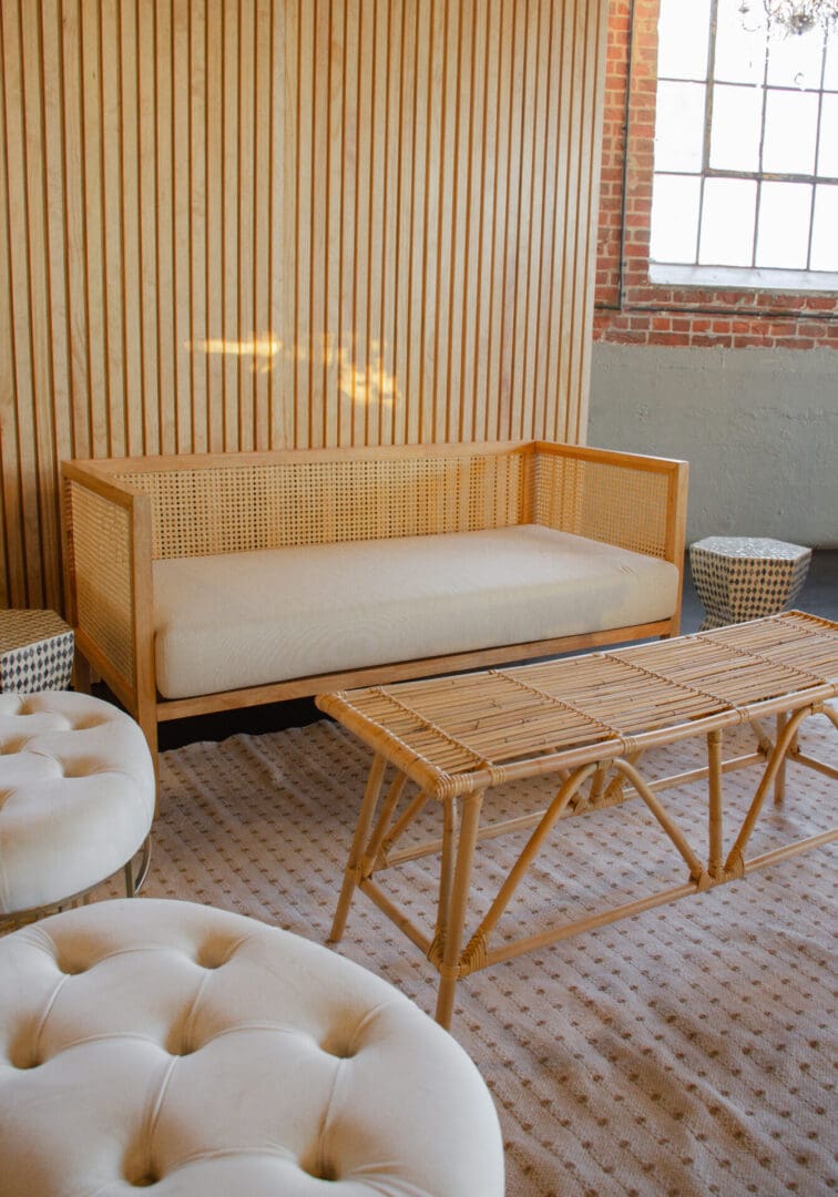 A minimalist living space featuring a rattan sofa, matching coffee table, tufted ottomans, and a textured rug, with a backdrop of vertical slat walls against a brick wall.