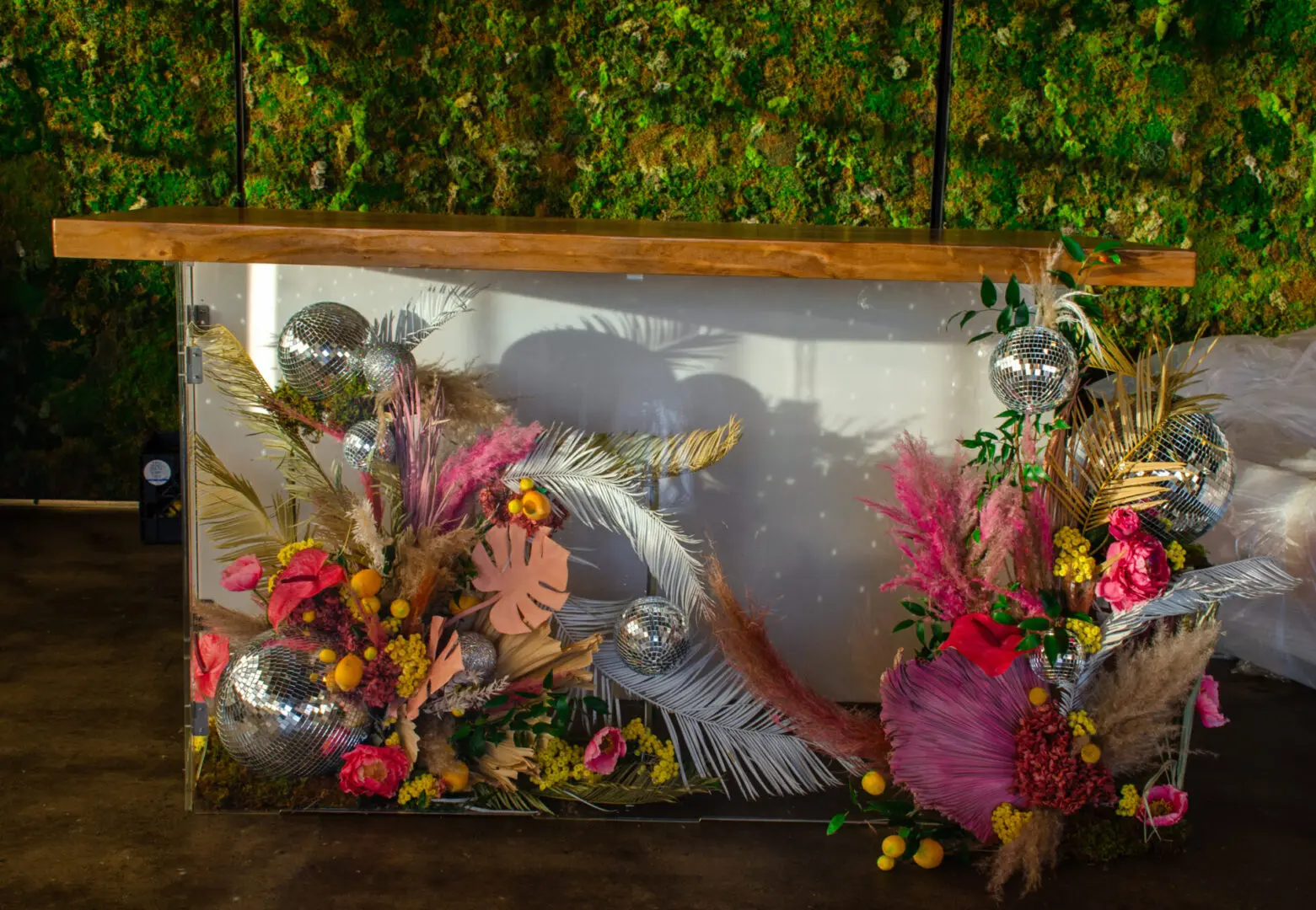 A decorative bar front adorned with colorful flowers, feathers, and disco balls, set against a backdrop of a greenery wall made for an event.