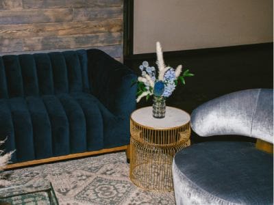 A cozy corner set up for an event, featuring a blue velvet sofa, a round wicker table with a floral arrangement, and a patterned rug.