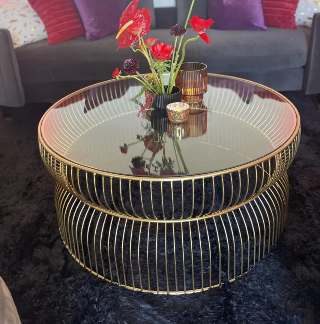 A round, gold-framed glass coffee table set for an event, with decorative items including red flowers in a vase and candles, placed on a black furry rug.