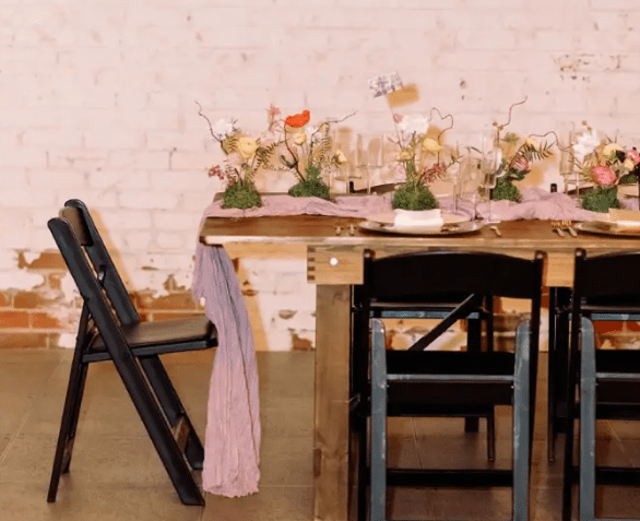 Event table setting with rustic floral centerpieces and folded chairs against an exposed brick wall.
