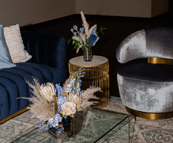 A cozy event sitting area with blue and gray velvet chairs, decorative throw pillows, and two arrangements of dried flowers on small tables.