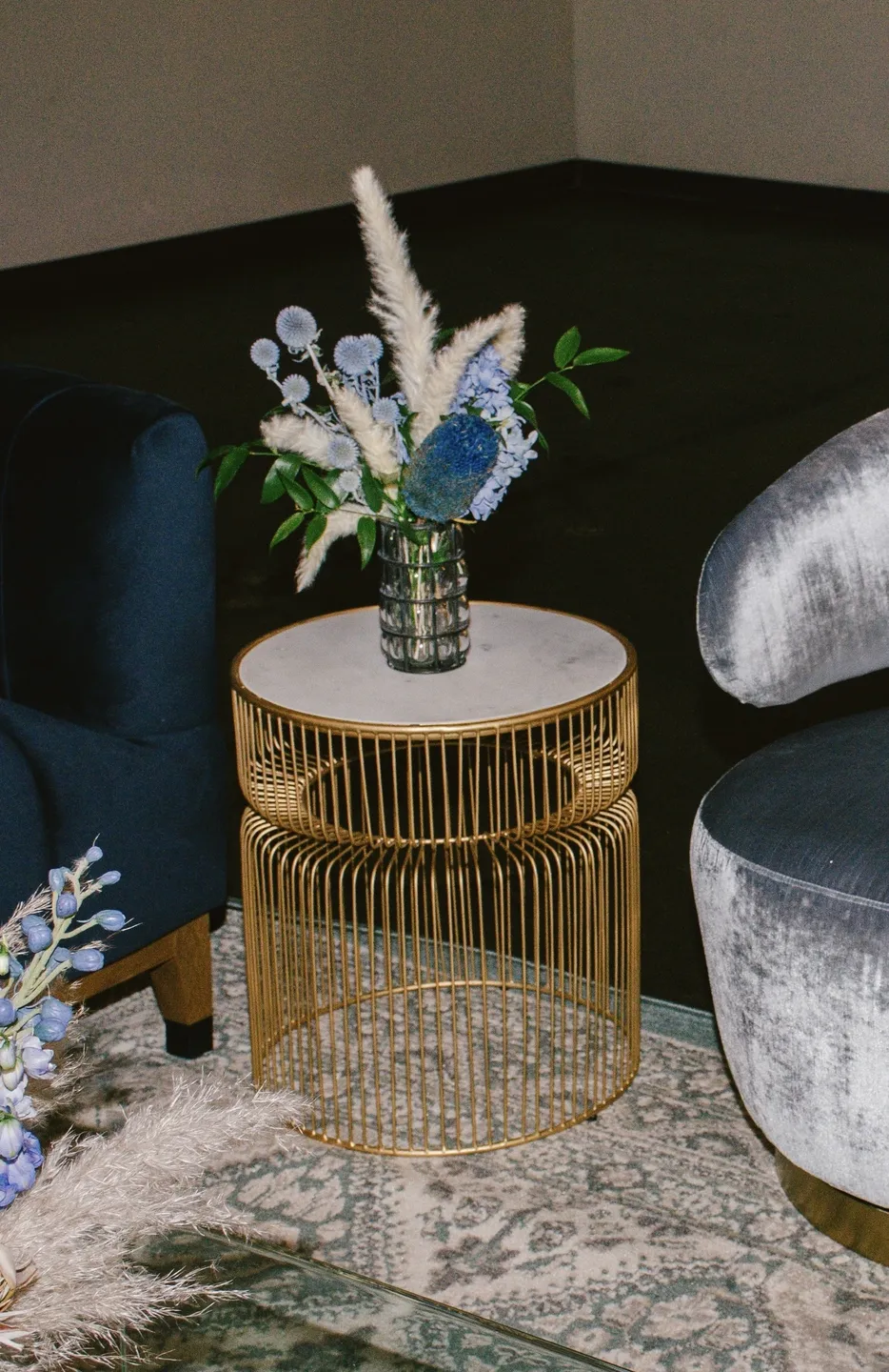 A modern living room prepared for an event, featuring a gold metal wire side table with a floral arrangement on top, flanked by two dark upholstered chairs.